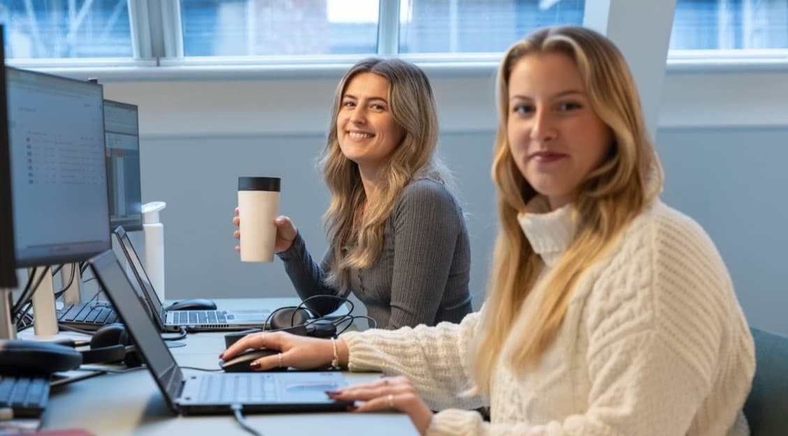 Two smiling females at work on their laptops looking at the camera.